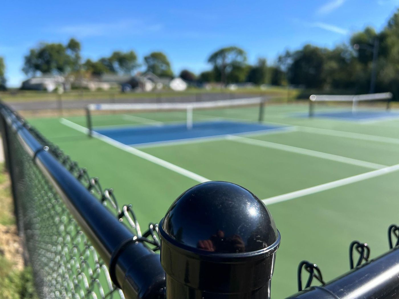 Tennis court viewed from a chain-link fence, with black fence post and blue sky above.