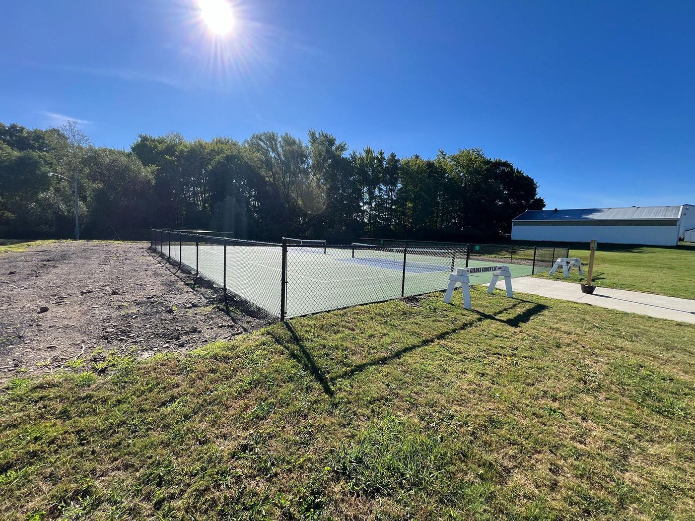 Outdoor tennis court by a grassy field under a bright blue sky, with a white building and trees nearby
