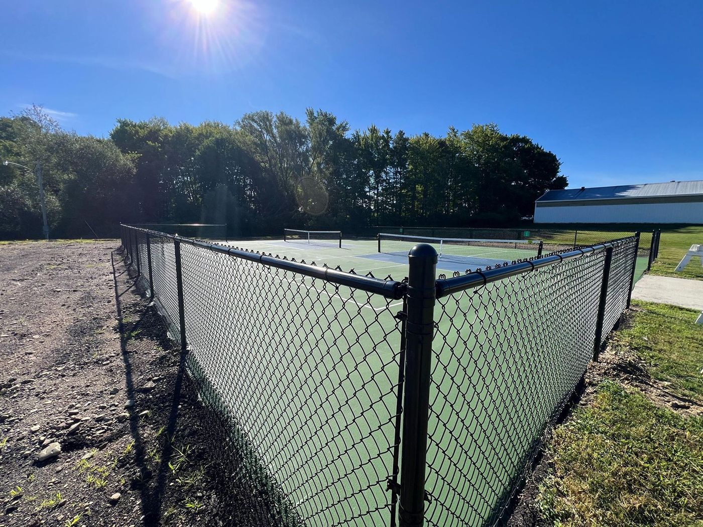 Chain-link fenced gravel enclosure beside a green field under a bright blue sky