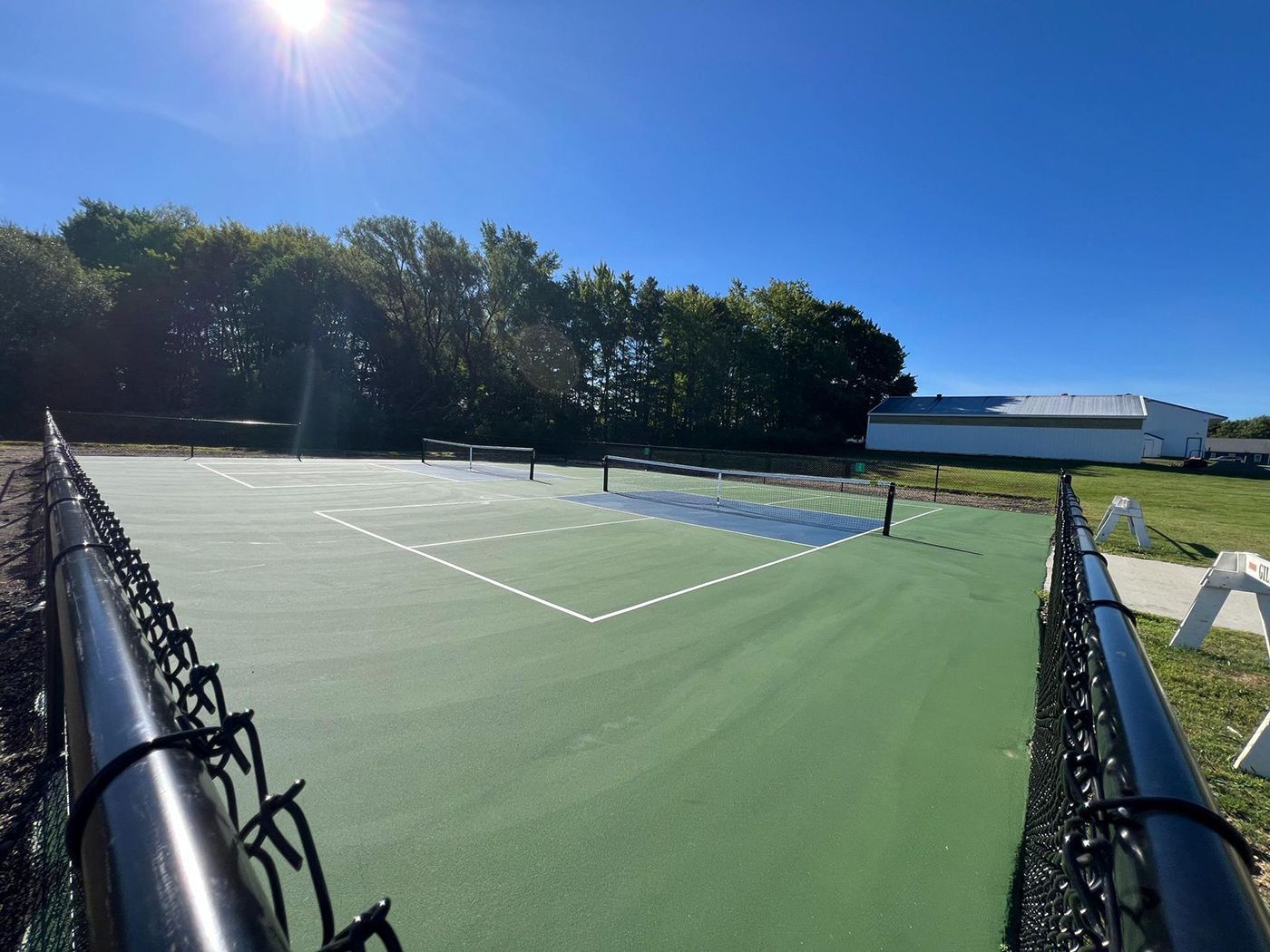 Tennis court under bright sun, viewed through a fence, with trees and buildings in the background.