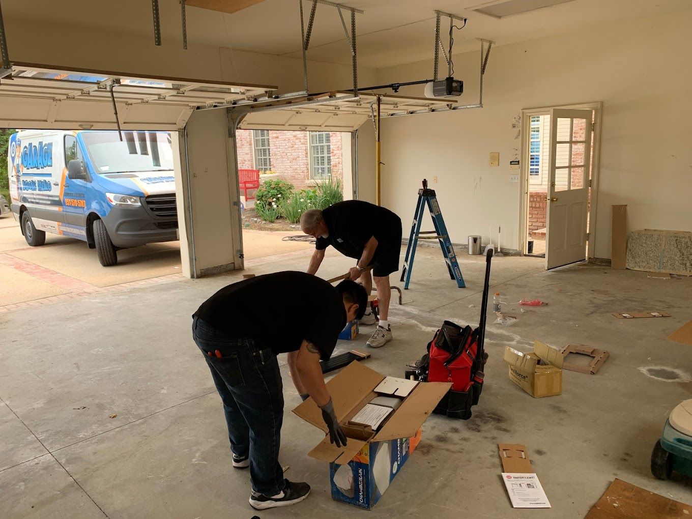 Two men are working in a garage with a van in the background.