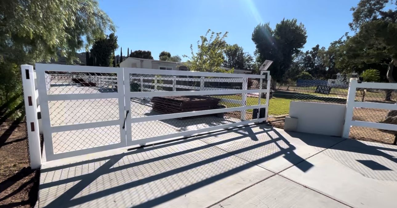 White gated entrance on a paved driveway with green grass and trees in the background, sunny day.