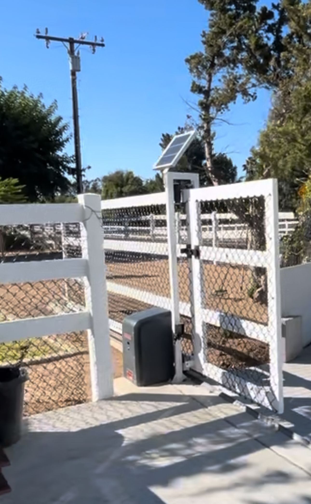 White gate with automatic opener, solar panel, and a wooden fence on a sunny day.