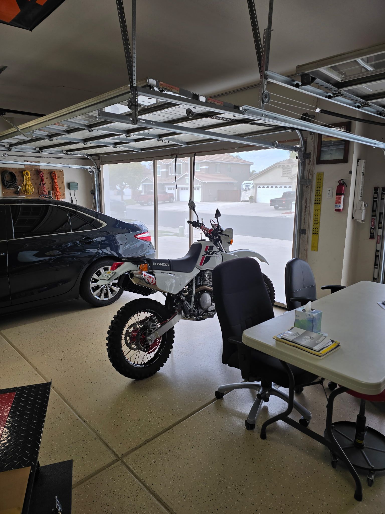 Motorcycle parked in garage, next to a car and table. Open garage door leads to street.