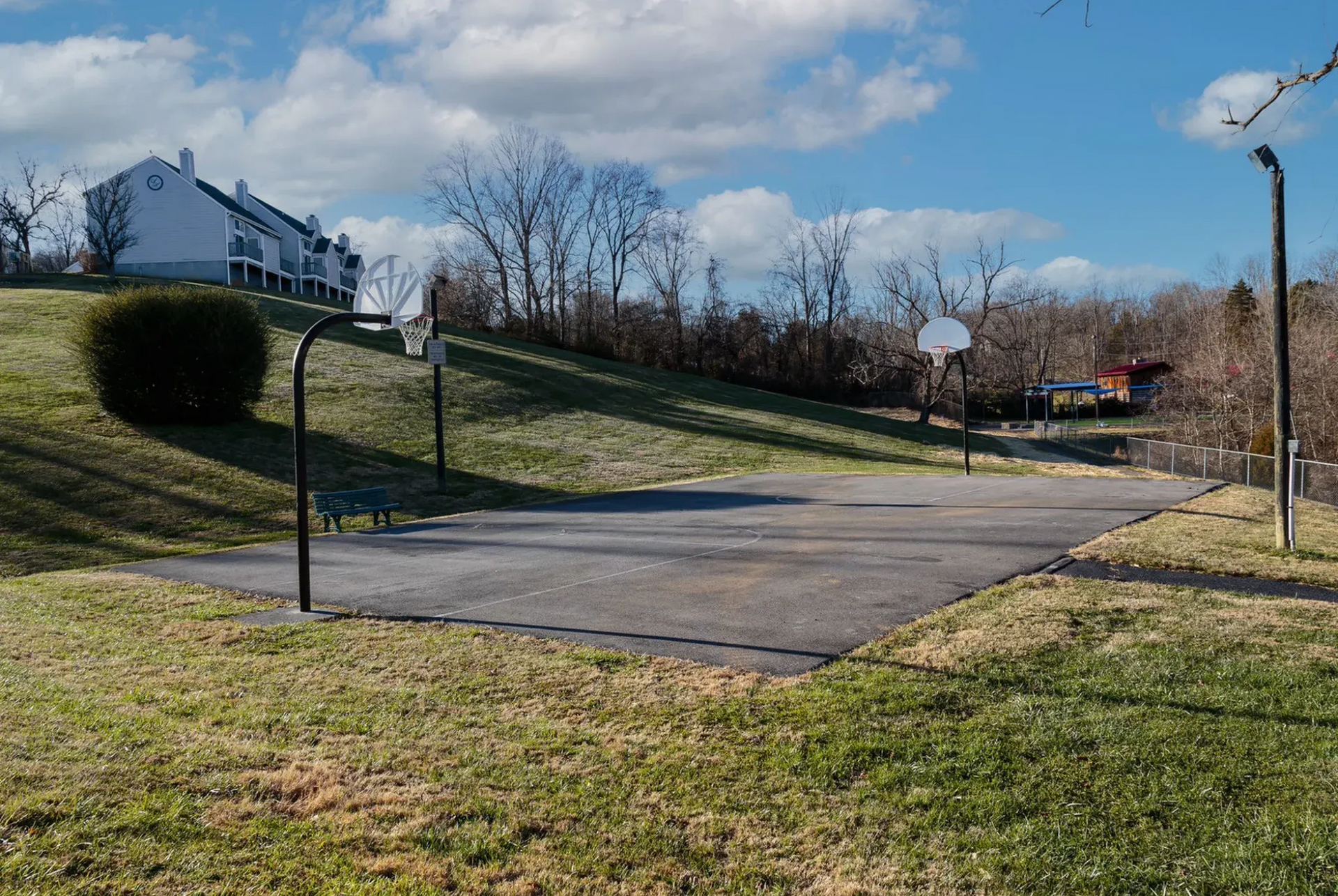 Outdoor basketball court with two hoops, set on a paved area near grassy hills and apartment buildings.