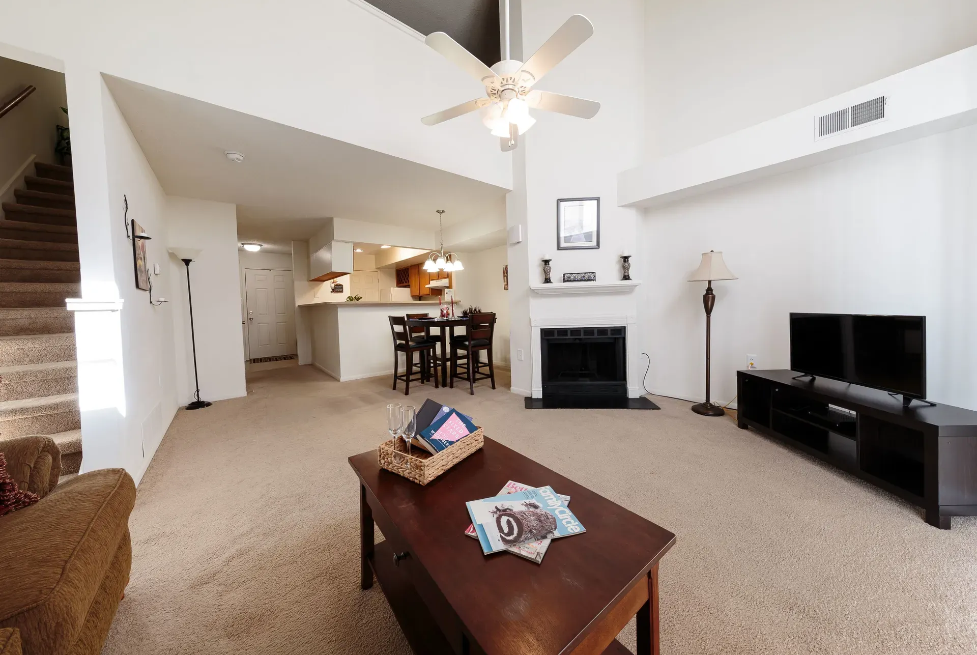 Open-concept living room with beige carpet, fireplace, TV stand, coffee table, and kitchen in the background.