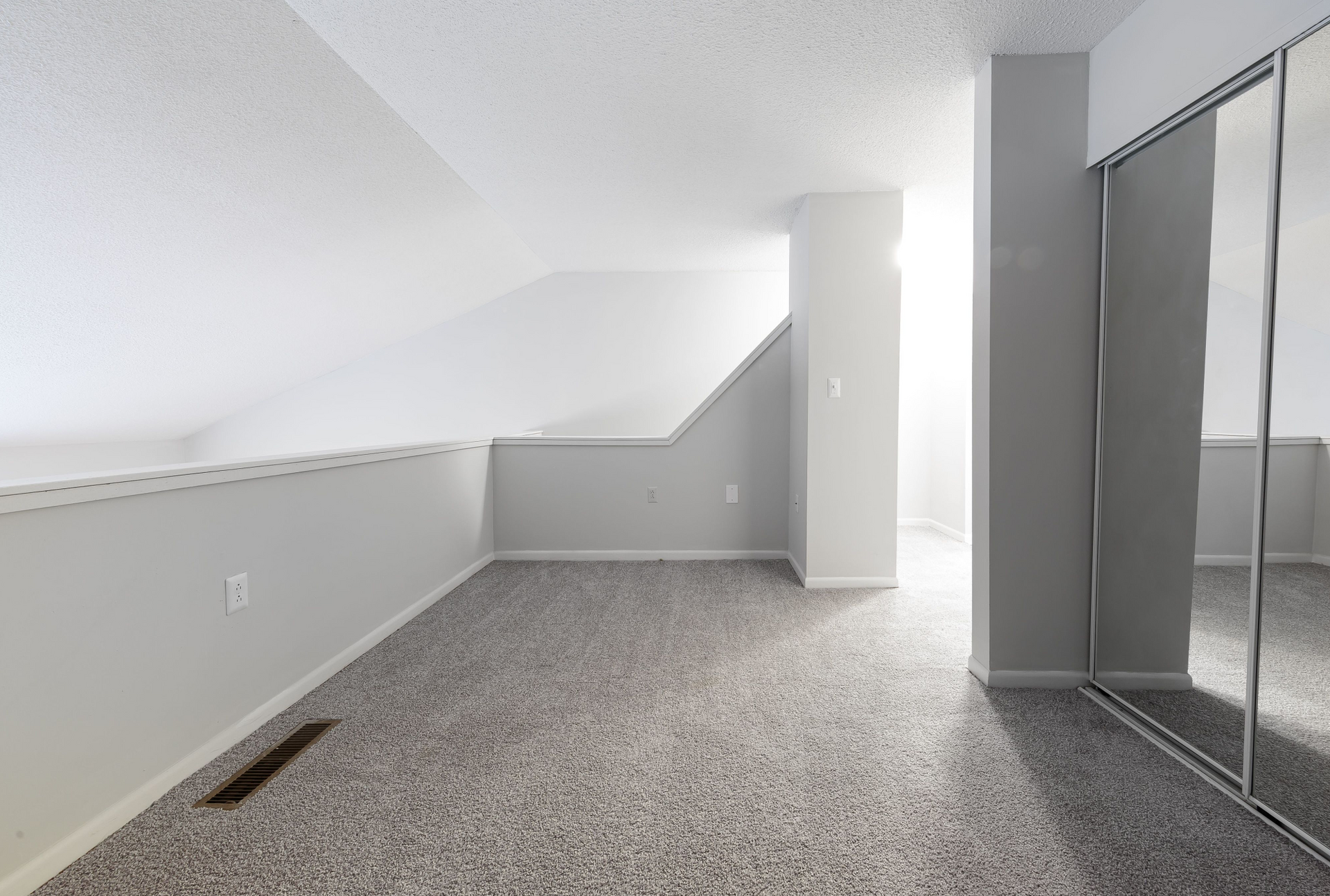 Interior view of a small attic-style room with sloped ceiling, carpet, and mirrored closet doors.