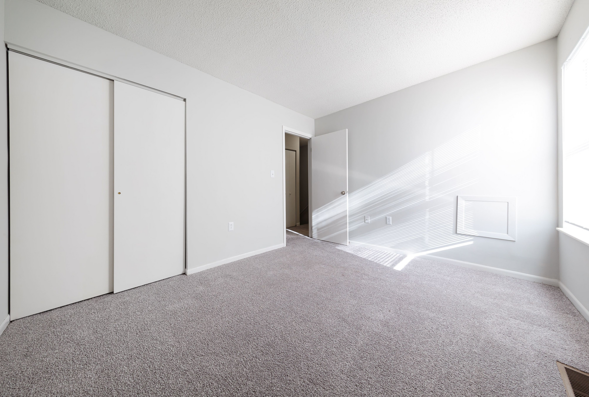Empty bedroom with sliding closet doors, open door, and sunlight on light gray walls.