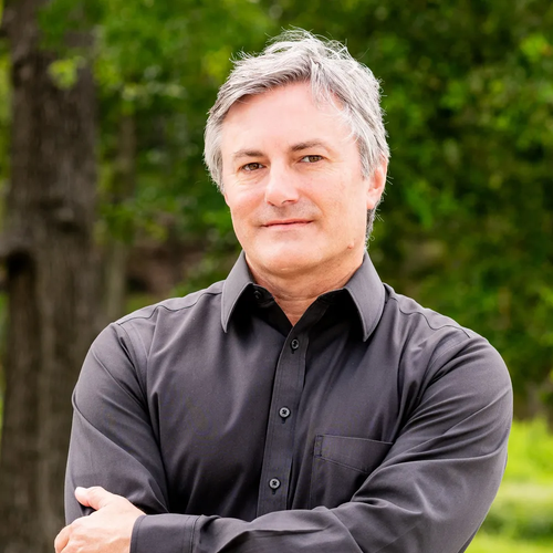 A person with grey hair wearing a black button-down shirt, standing with arms crossed against a blurred green background.
