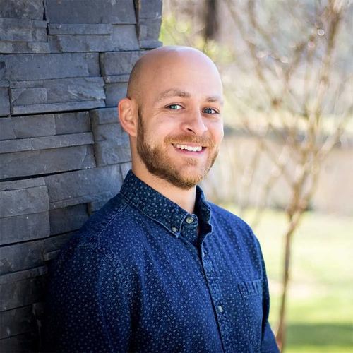 A smiling person with a beard and a shaved head, wearing a blue patterned button-down shirt, leans against a stone wall.
