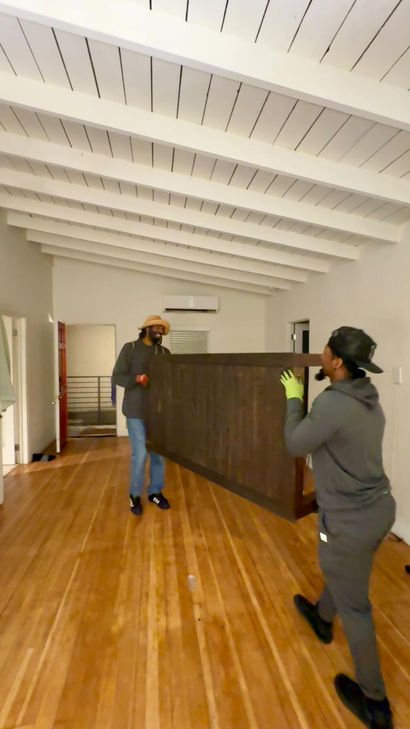 Two people moving a large dark divider in a bright room with wood floors.