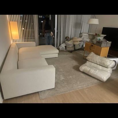 Living room with white sectional sofa, beige rug, lamp, and stacked moving items near the window