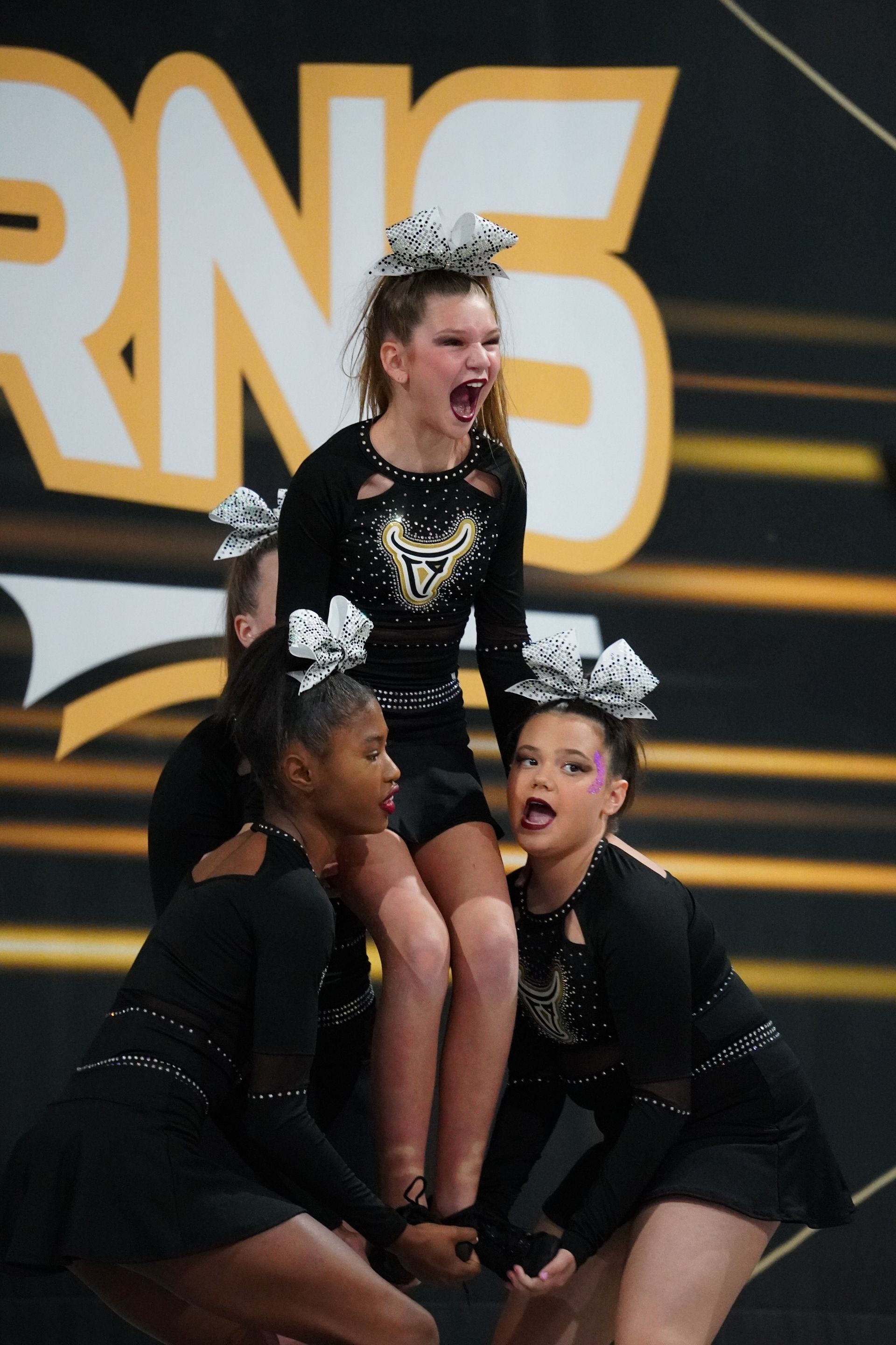 Cheerleaders in black and gold uniforms performing a pyramid. Girl on top yells. Indoor competition.