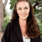 Portrait of a smiling woman with long curly hair wearing a dark jacket outdoors, blurred trees behind her