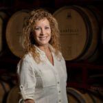 Woman in a white blouse standing in front of wooden barrels, smiling in a dim cellar.