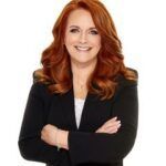 Smiling person with red hair in a black blazer, arms crossed against a white background