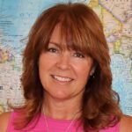 Smiling woman with auburn hair in a pink top, standing before a colorful map background