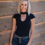 Woman in a black top and jeans standing against a wooden wall, smiling at the camera.
