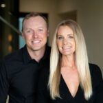 Two people smiling indoors, one in a black shirt and one in a black blazer, against a softly blurred background