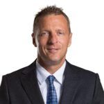 Studio headshot of a man in a dark suit, white shirt, and blue tie on a white background