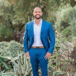 Man in a blue suit smiling outdoors among greenery, with hands in his pockets.