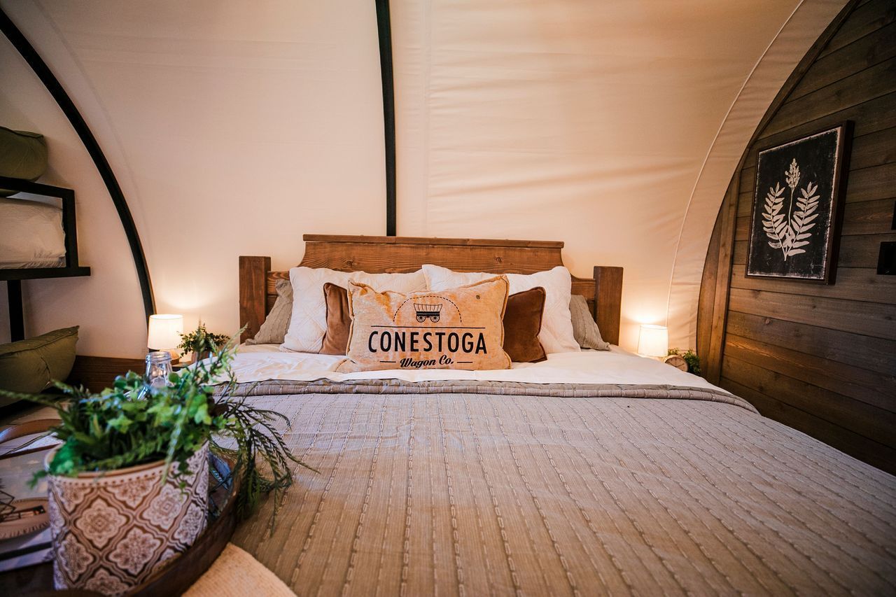 A cozy bedroom inside a Conestoga wagon glamping unit at Clay Cosmos showing a bed with a wooden headboard and pillows