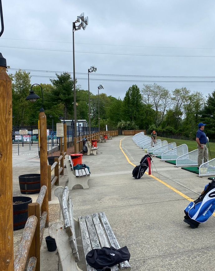 Golf driving range with benches, bags, and people. Gray concrete path, yellow lines, and lighting poles.