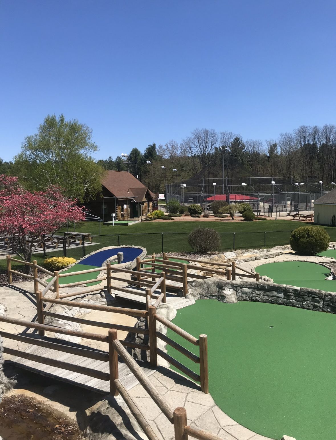 Miniature golf course with green fairways, wooden bridges, and a building on a sunny day.