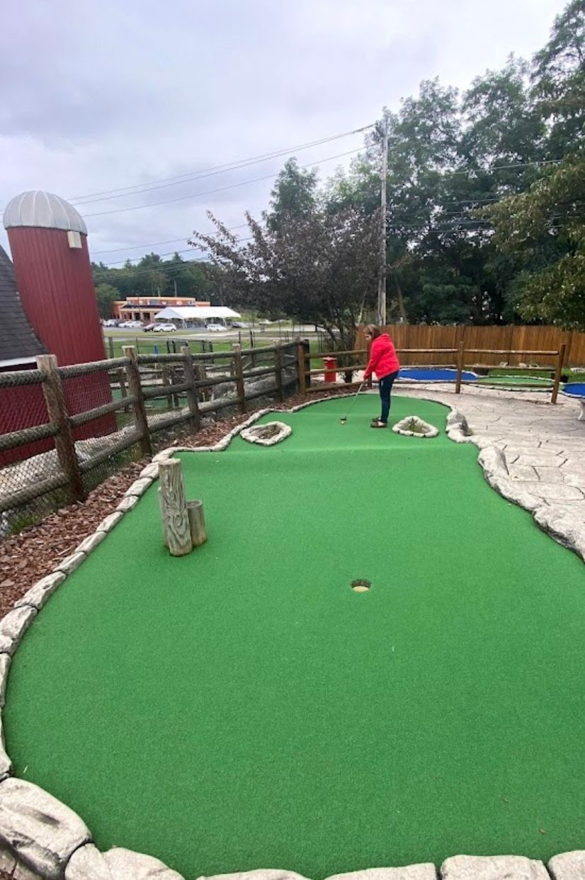 Person playing miniature golf on a green course with a red barn in the background.