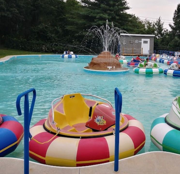 Bumper boats on a pool with a fountain feature. Boats are red, yellow, and blue. Cloudy day.