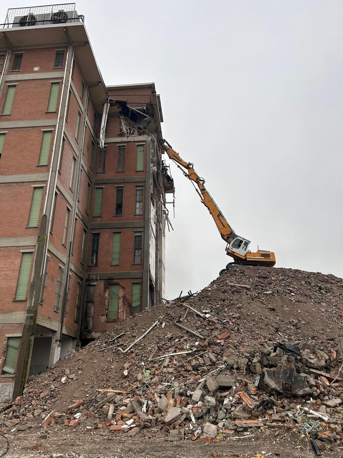 Un escavatore giallo demolisce un edificio in mattoni sotto un cielo azzurro e limpido.