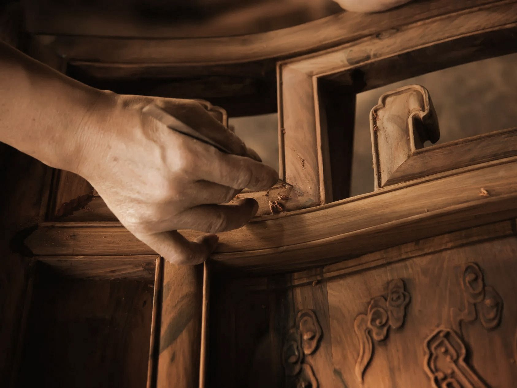 Hand touches wooden furniture, examining detail. Interior shot with carvings and frame.