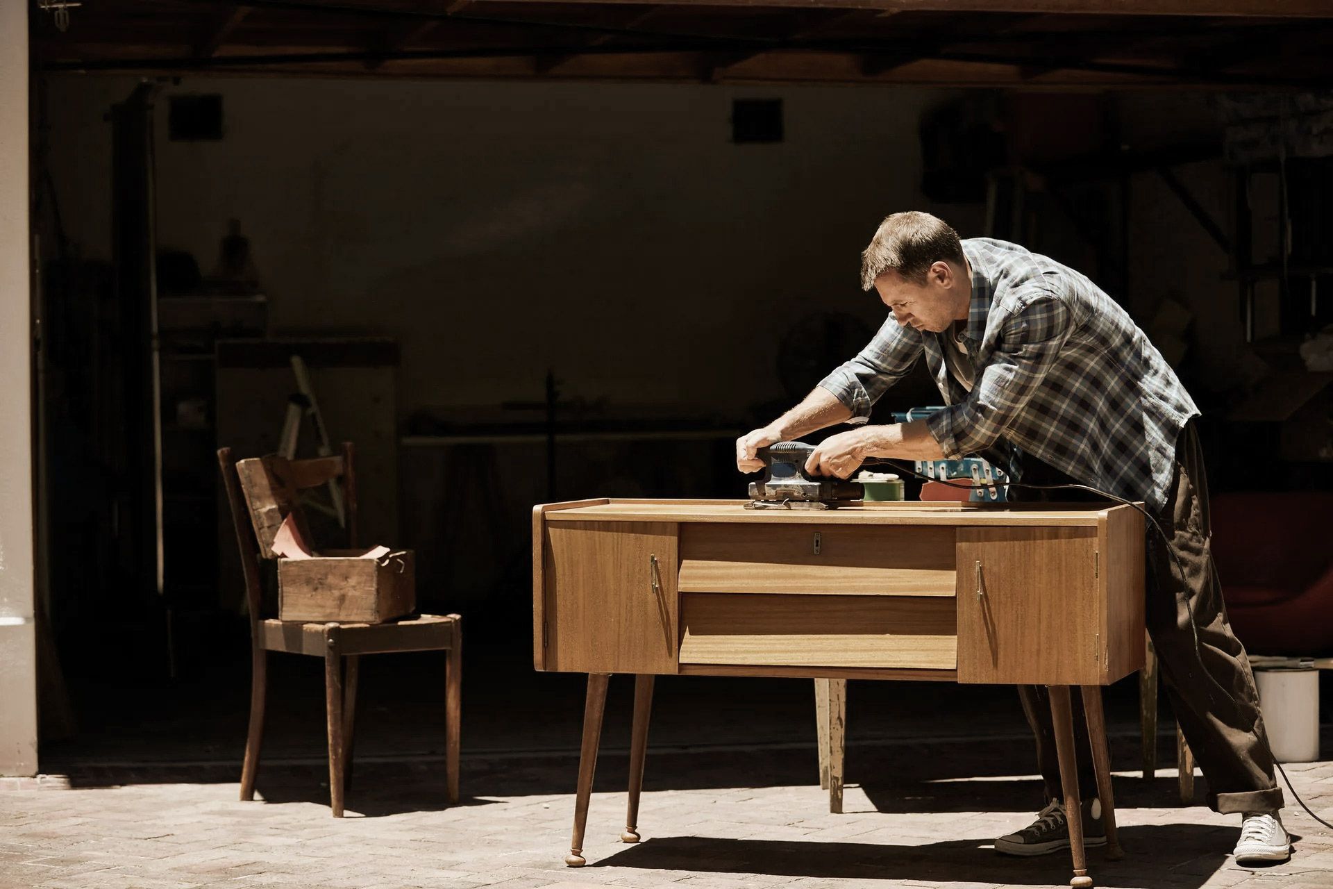 Man sands a wooden cabinet in a garage; a toolbox sits on a chair.