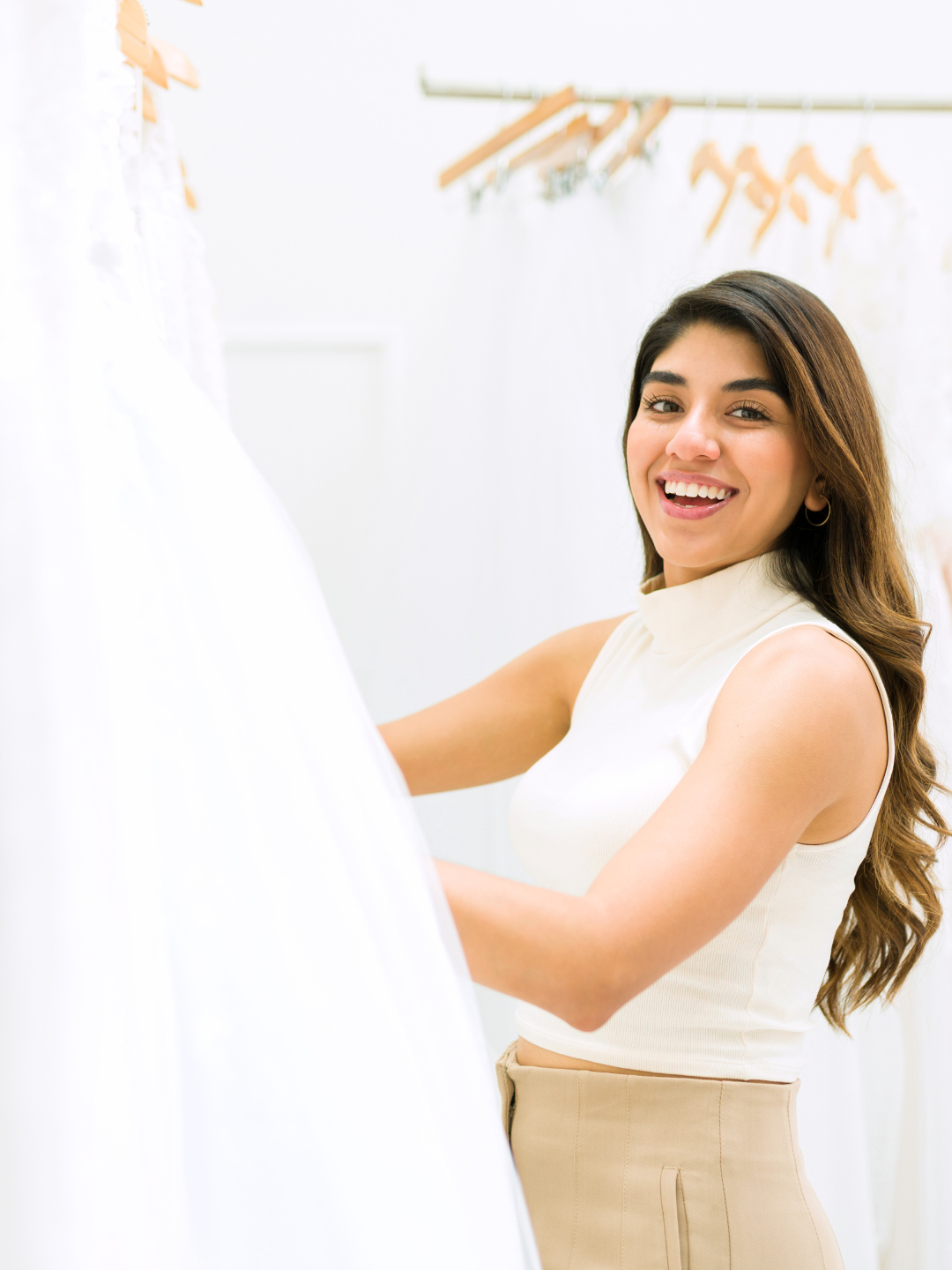 Woman smiling, touching a white wedding dress in a bridal shop.