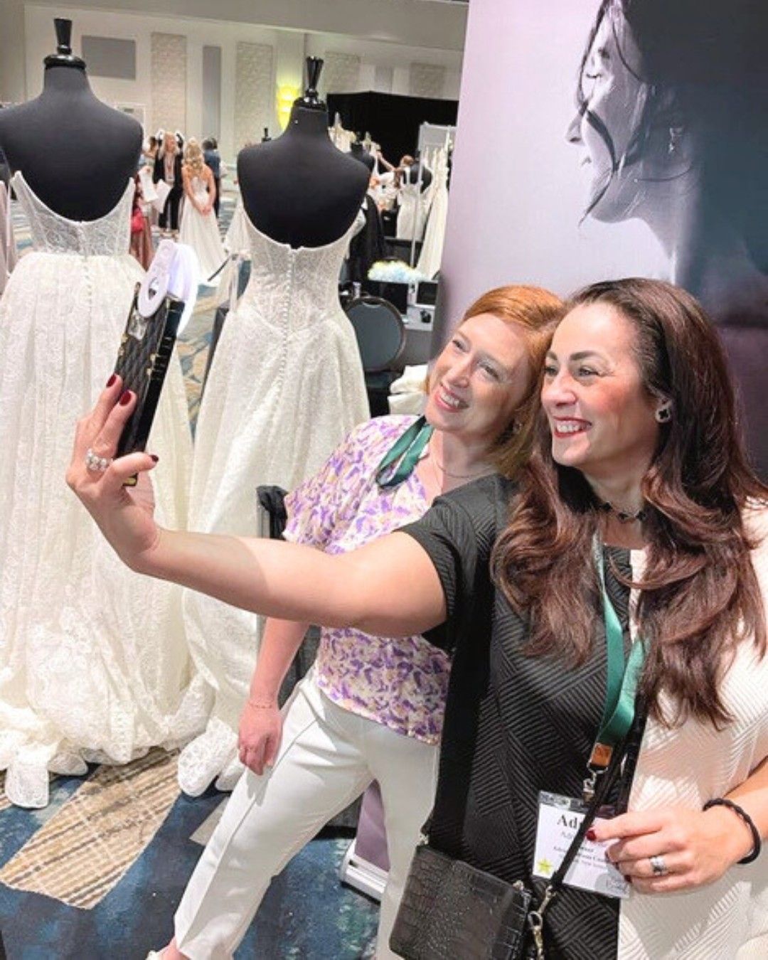 Two women take a selfie in a bridal shop, dresses on mannequins in the background.