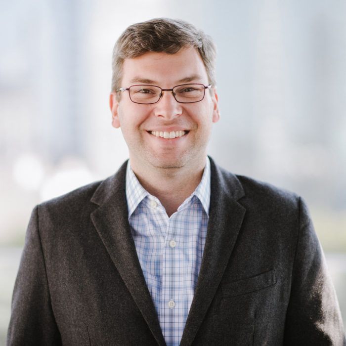 Man with glasses smiles, wearing a blazer over a blue-checkered shirt in a bright setting.
