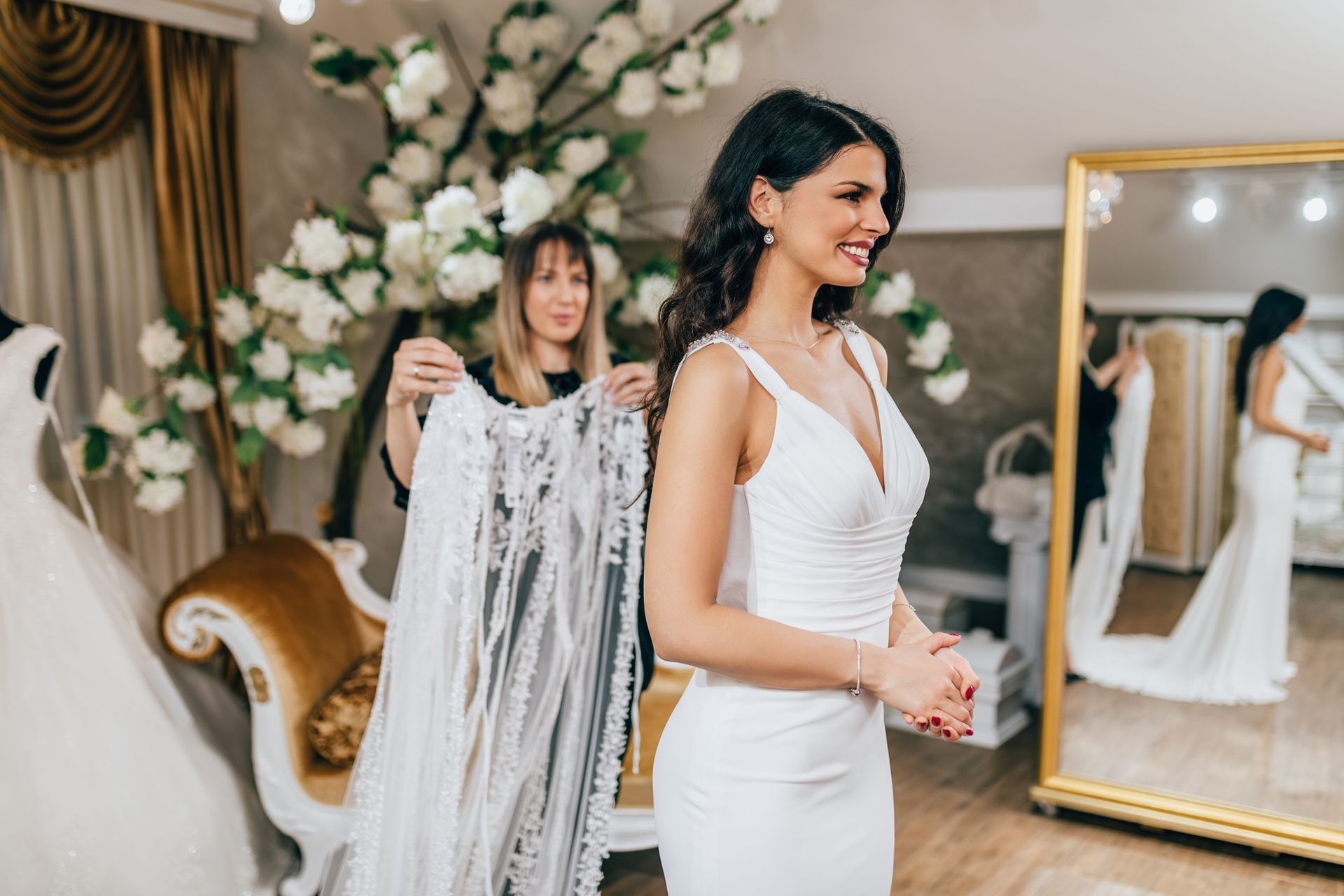 Woman in wedding dress in a bridal shop; shop assistant holds a lace veil, smiling.