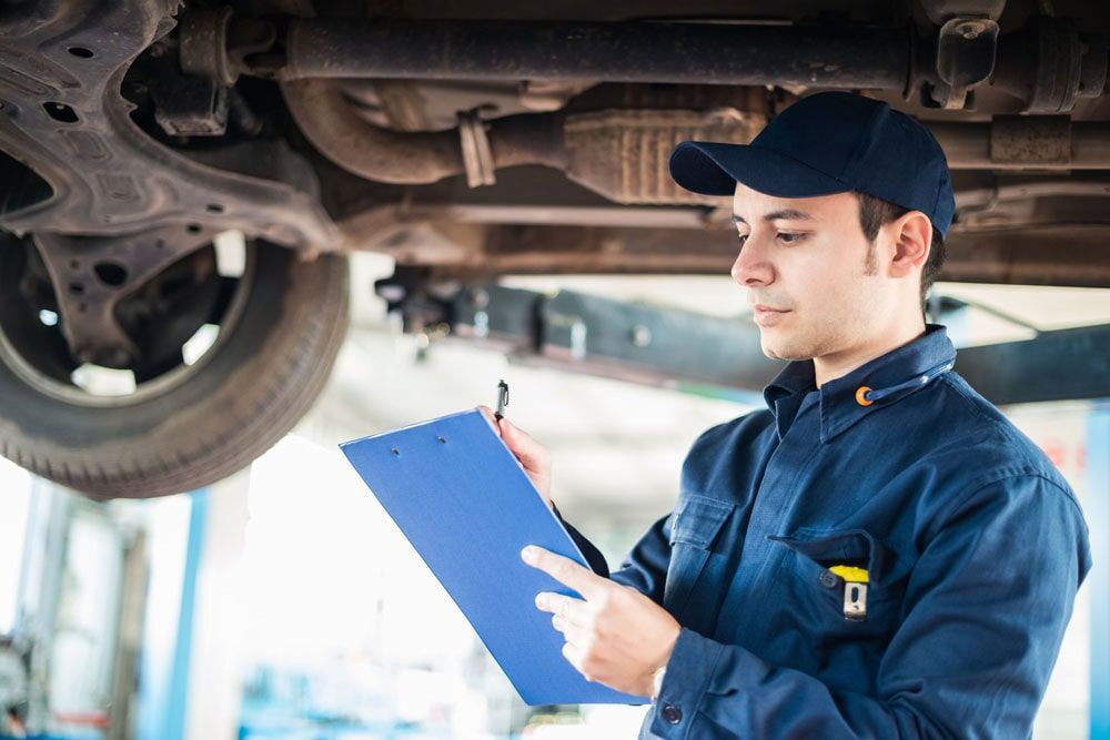 Mechanic in Blue Uniform Inspects Undercarriage of Car, Holding Clipboard — Peter Mac's Mechanical in Morisset, NSW