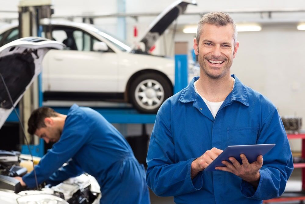 Mechanic Smiling at Camera Holding Tablet, Another Working on Car in Garage — Peter Mac's Mechanical in Morisset, NSW