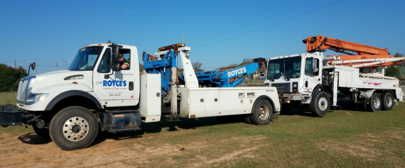 A white tow truck towing a white concrete pump truck on a grassy field under a blue sky.