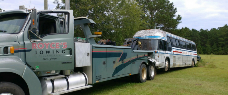 Tow truck towing a bus in a grassy field. The truck is green, the bus is white. Royce's Towing logo on truck.
