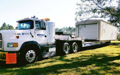 White semi-truck transporting a small white modular home on a flatbed trailer in a grassy area.
