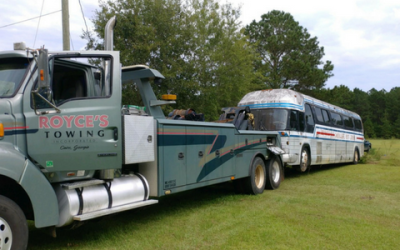 Tow truck towing a bus on a grassy field; the truck is teal, and the bus is white.