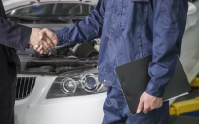 Mechanic in blue jumpsuit shakes hands with customer near a car with the hood open.