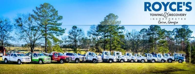 Row of tow trucks on a grassy field with trees under a blue sky. Royce's Towing logo in the top right.