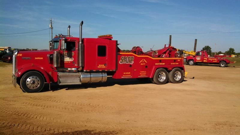 Red tow trucks on a dirt lot under a blue sky, one is a large semi-truck.