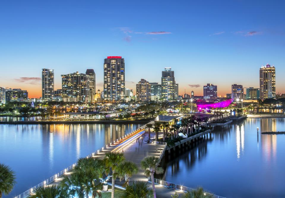 City skyline at dusk with lights reflecting on water; pier and palm trees in foreground.