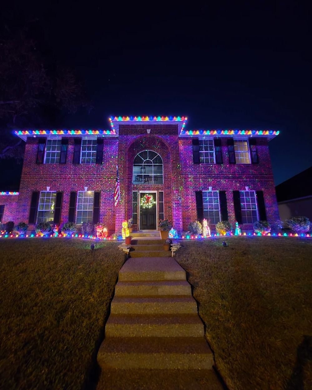 Brick house lit with colorful Christmas lights at night, with a walkway leading to the front door.