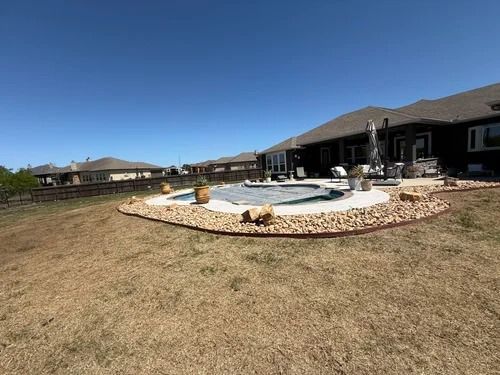Backyard patio with a small pool and stone border beside a single-story house under a blue sky