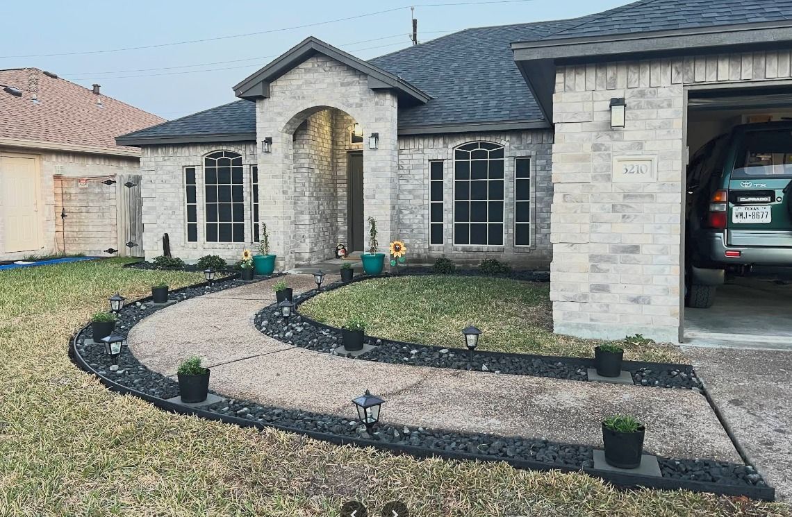 Stone house with curved gravel front path, black edging, small potted plants, and an open garage with car.